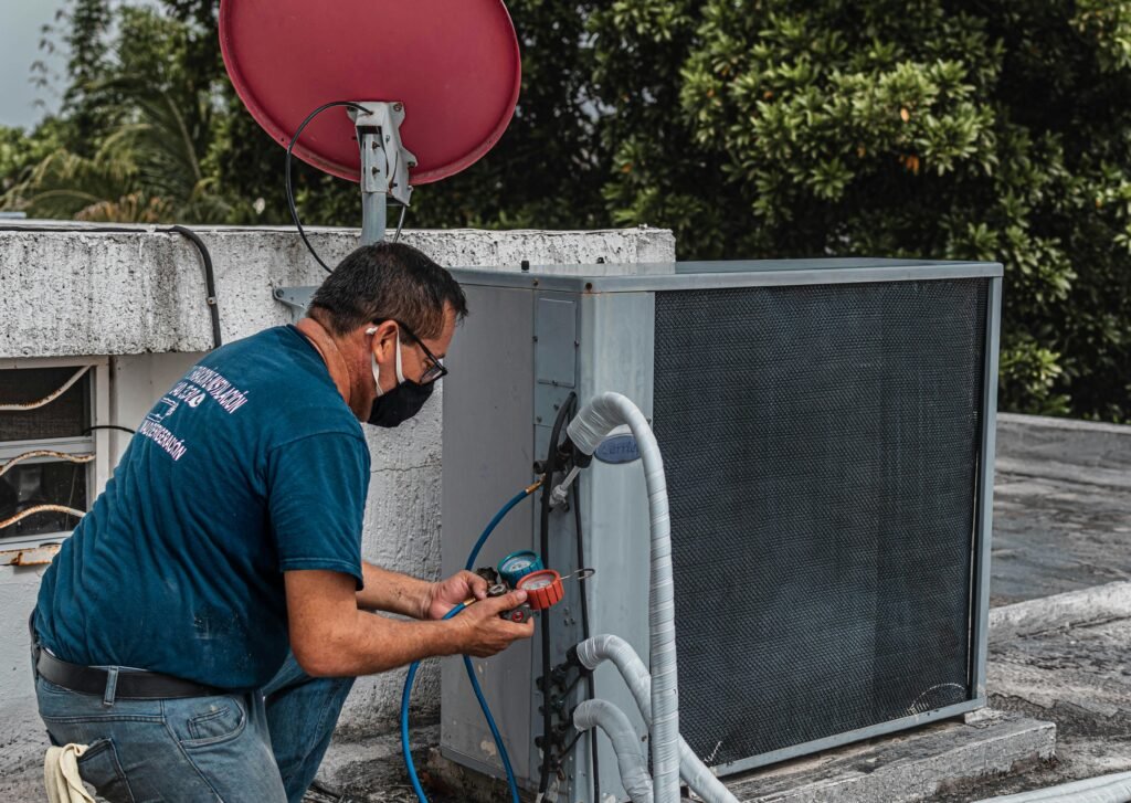 pexels-photo-5463575-5463575 Technician repairing an air conditioner unit outdoors, wearing a facemask and using a manifold gauge.